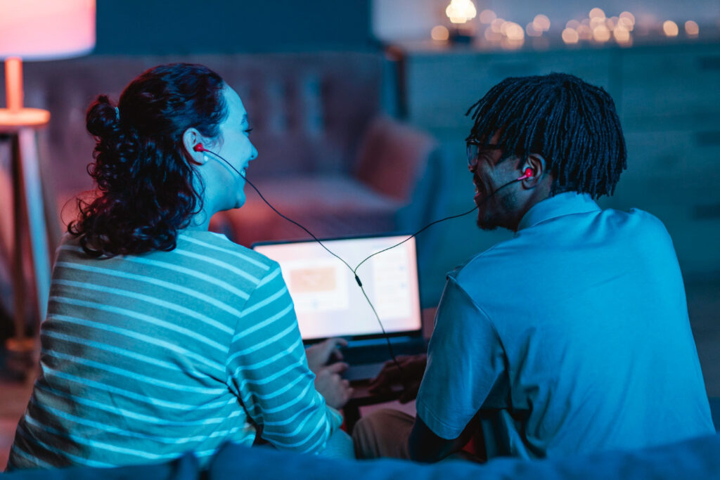 A couple seen from behind, sitting at home using a laptop and earphones.